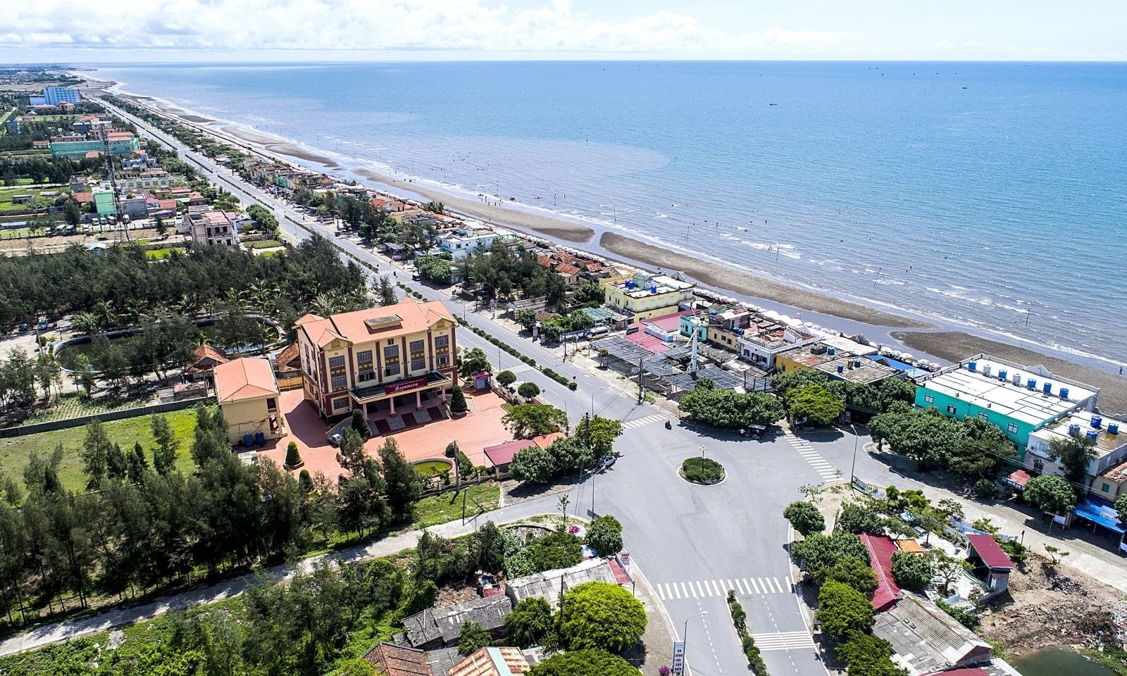 Ảnh flycam thị trấn ven biển Ninh Bình: con đường thẳng chạy dọc bờ biển, tòa nhà ngói đỏ giữa hàng phi lao, bãi cát và biển xanh phía xa